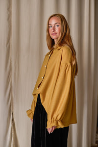 A woman with long, reddish hair stands against a beige curtain backdrop. She is wearing a mustard-yellow Poet Shirt | Japanese Wool Gauze Gold with billowy sleeves and black pants. She gazes calmly at the camera with a relaxed posture.