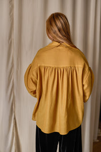 A person with long reddish-blonde hair is standing with their back to the camera, wearing a loose-fitting mustard yellow Poet Shirt | Japanese Wool Gauze Gold and black pants. The background features light-colored curtains.