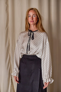 A woman with long blonde hair wears a white Ribbon Shirt | White Polkadot Silk Charmeuse adorned with black polka dots and a black ribbon tie at the neck. She pairs it with a high-waisted, black skirt. The background is a soft beige curtain. She stands facing the camera with a neutral expression.