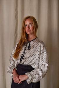 A woman with long, strawberry-blonde hair stands in front of a light-colored curtain. She is wearing a Ribbon Shirt | White Polkadot Silk Charmeuse and a black skirt. She holds her hands together in front of her and gazes at the camera.