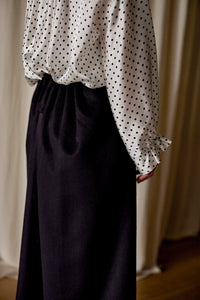 A person is wearing a white blouse with black polka dots, featuring ruffled cuffs, and a Layered Skirt | Cashmere. Handmade in San Francisco, the photo captures the torso and part of the legs against a neutral backdrop of beige curtains.