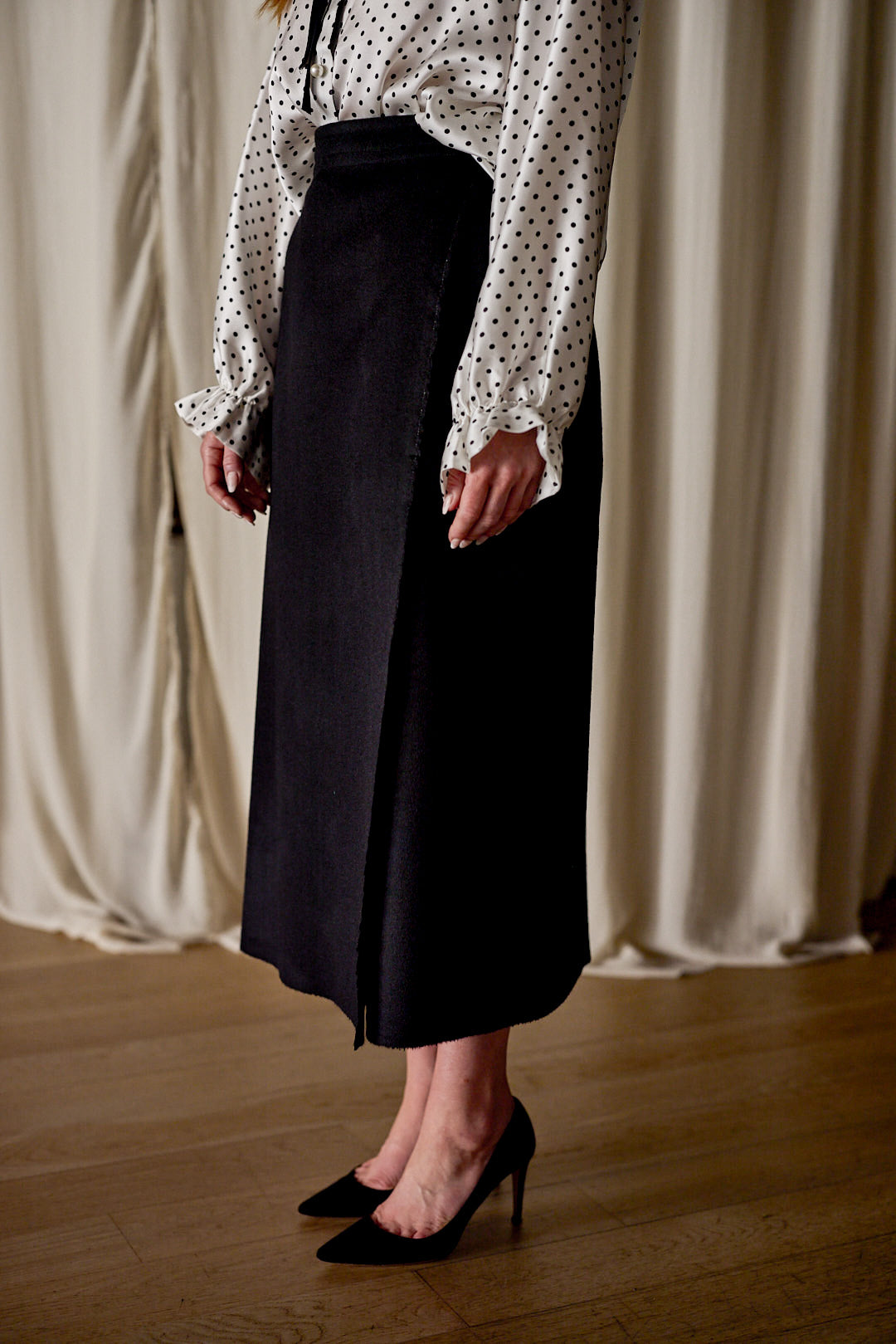 Person standing on wooden floor, wearing a white long-sleeve polka dot blouse with ruffled cuffs, paired with a high-waisted Layered Skirt | Cashmere featuring an asymmetrical front seam. Black high-heeled shoes complete the look against a background of light-colored curtains.