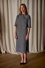 Load image into Gallery viewer, A woman with long, light brown hair wearing a knee-length, gray Qipao Dress | Japanese Wool Gauze Houndstooth and black shoes is standing on a wooden floor in front of cream-colored curtains, looking at the camera. The dress embodies sustainable design through its use of Japanese wool gauze.