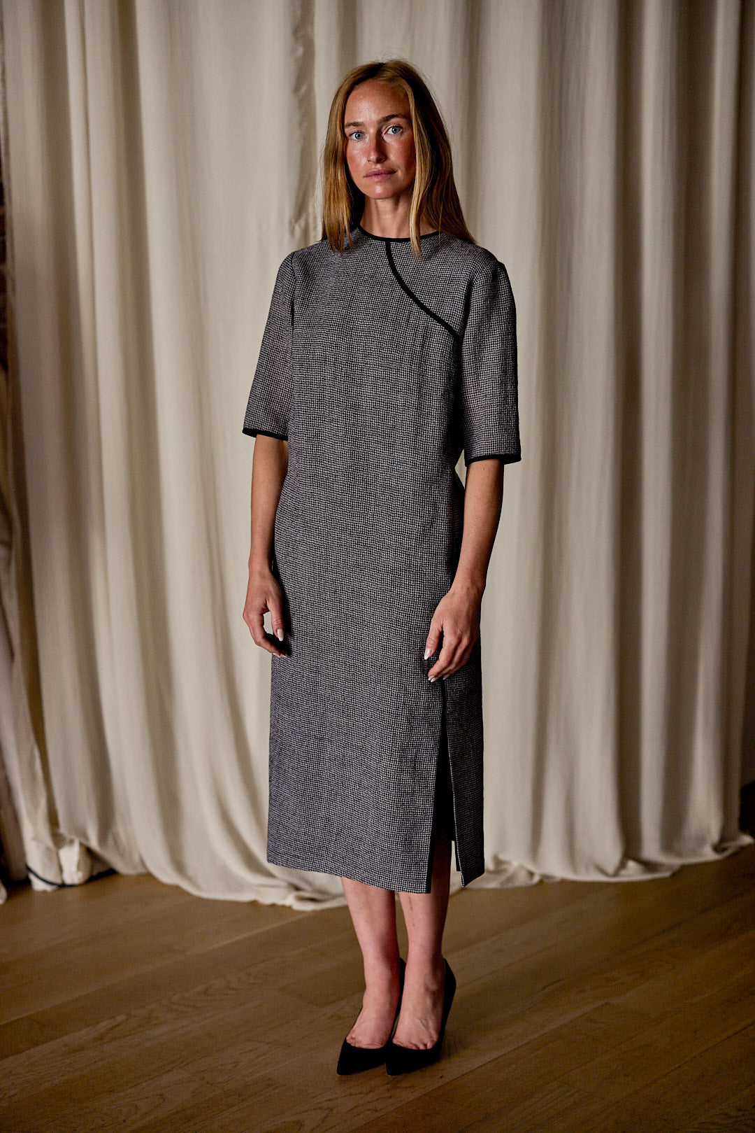 A woman with long, light brown hair wearing a knee-length, gray Qipao Dress | Japanese Wool Gauze Houndstooth and black shoes is standing on a wooden floor in front of cream-colored curtains, looking at the camera. The dress embodies sustainable design through its use of Japanese wool gauze.