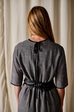 Load image into Gallery viewer, A person with long blonde hair is standing with their back facing the camera. They are wearing a Qipao Dress | Japanese Wool Gauze Houndstooth, featuring a black ribbon tied at the neck and a wide black belt around the waist. The background consists of beige curtains.