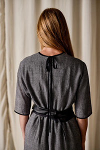 A person with long blonde hair is standing with their back facing the camera. They are wearing a Qipao Dress | Japanese Wool Gauze Houndstooth, featuring a black ribbon tied at the neck and a wide black belt around the waist. The background consists of beige curtains.
