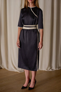 A woman stands in a room with draped curtains, wearing a Qipao Dress | Silk Charmeuse Black with white trim accents along the neckline, waist, and edges. The dress has elbow-length sleeves reminiscent of a Qipao dress, and she is wearing black pointed-toe heels. Her hands are relaxed by her sides.