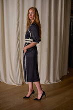 Load image into Gallery viewer, A woman with long, light brown hair stands on a wooden floor in front of white curtains. She is wearing a dark knee-length Qipao Dress | Silk Charmeuse Black with a white belt and trim, made from sustainable design principles, paired with black high-heeled shoes. Her hands are in her pockets, and she looks at the camera with a slight smile.