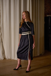 A woman with long red hair stands in front of a curtain, wearing a Qipao Dress | Silk Charmeuse Black with white trim and a side slit. She has black high heels on and is gazing to her right. The background features a wooden floor and partially visible dark furniture.