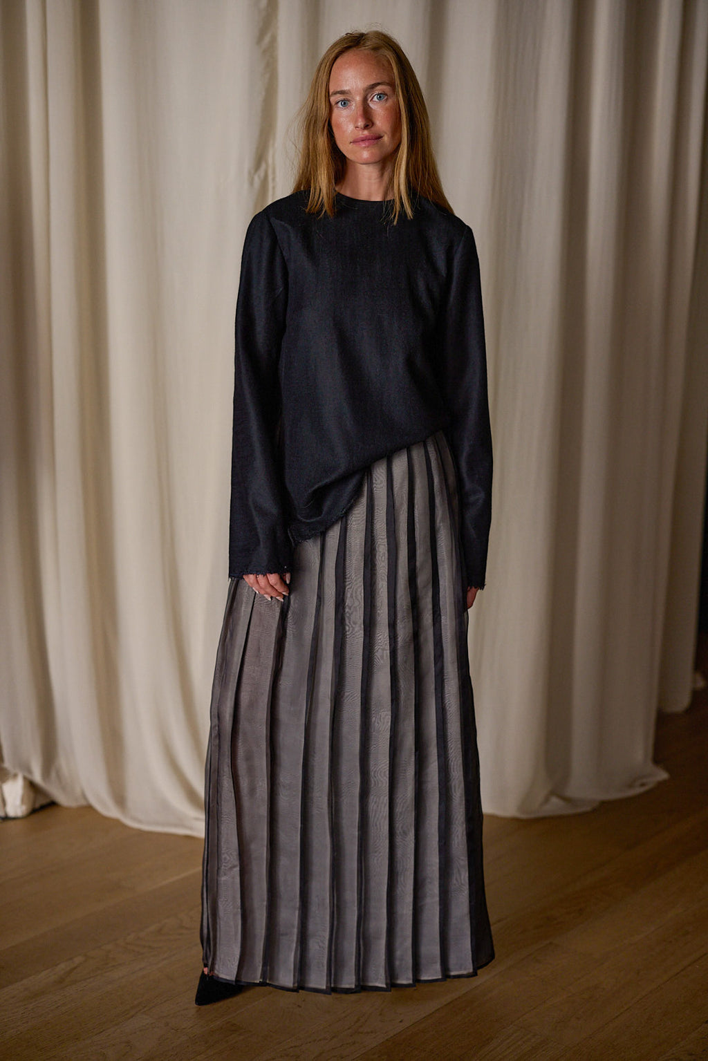 A person with long hair stands indoors in front of a light-colored curtain, wearing a dark long-sleeved top and an Atelier Lan Jaenicke Hand Pleated Wrap Skirt | Silk Organza with vertical stripes. The flooring is wooden.