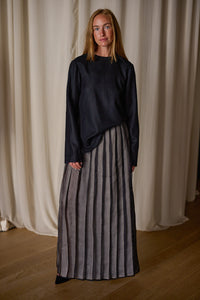 A person stands in front of draped curtains, wearing a Crewneck shirt - Japanese Wool Gauze | Black and a floor-length gray skirt with vertical black stripes. Crafted from Japanese wool gauze, this handmade ensemble complements their long blonde hair and neutral expression against the light-colored background and wooden floor.