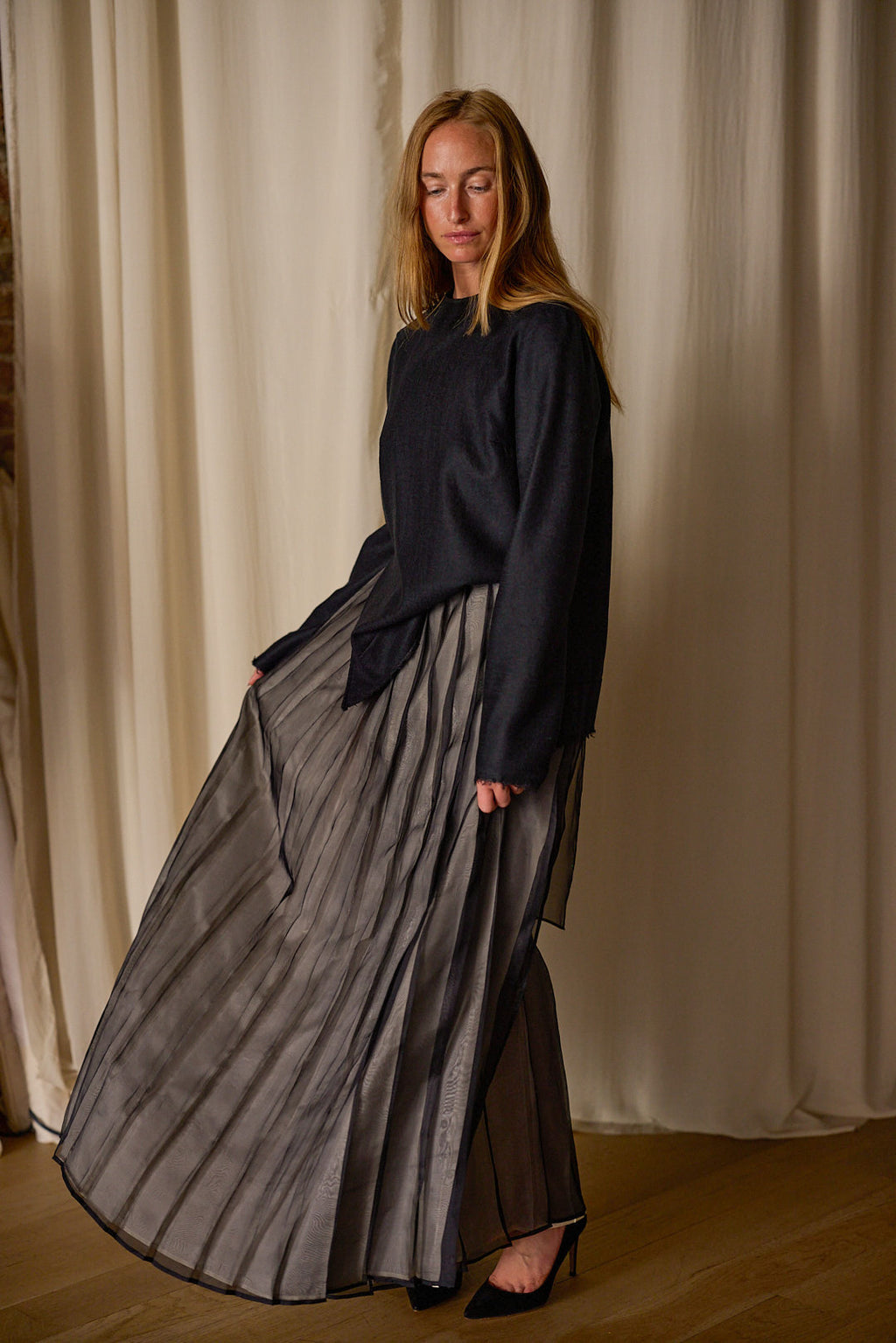 A woman with long, blonde hair stands in front of a light-colored curtain. She is wearing a dark, long-sleeved top and a flowing, pleated gray Hand Pleated Wrap Skirt | Silk Organza by Atelier Lan Jaenicke. Her eyes are closed, and she stands with one foot slightly in front of the other, creating a serene and elegant pose.
