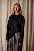 A woman with long, straight hair stands against a neutral curtain backdrop, wearing a Crewneck shirt - Japanese Wool Gauze | Black and a gray pleated skirt crafted from Japanese wool gauze. She looks directly at the camera with a calm expression, embodying timeless sustainable design.