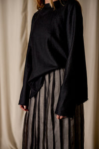 A person standing against a beige curtain background is wearing a Crewneck shirt - Japanese Wool Gauze | Black and a high-waisted, ankle-length, pleated skirt made from Japanese wool gauze with black and grey vertical stripes. The focus is on the sustainable design of the clothing, without showing the full face of the person.