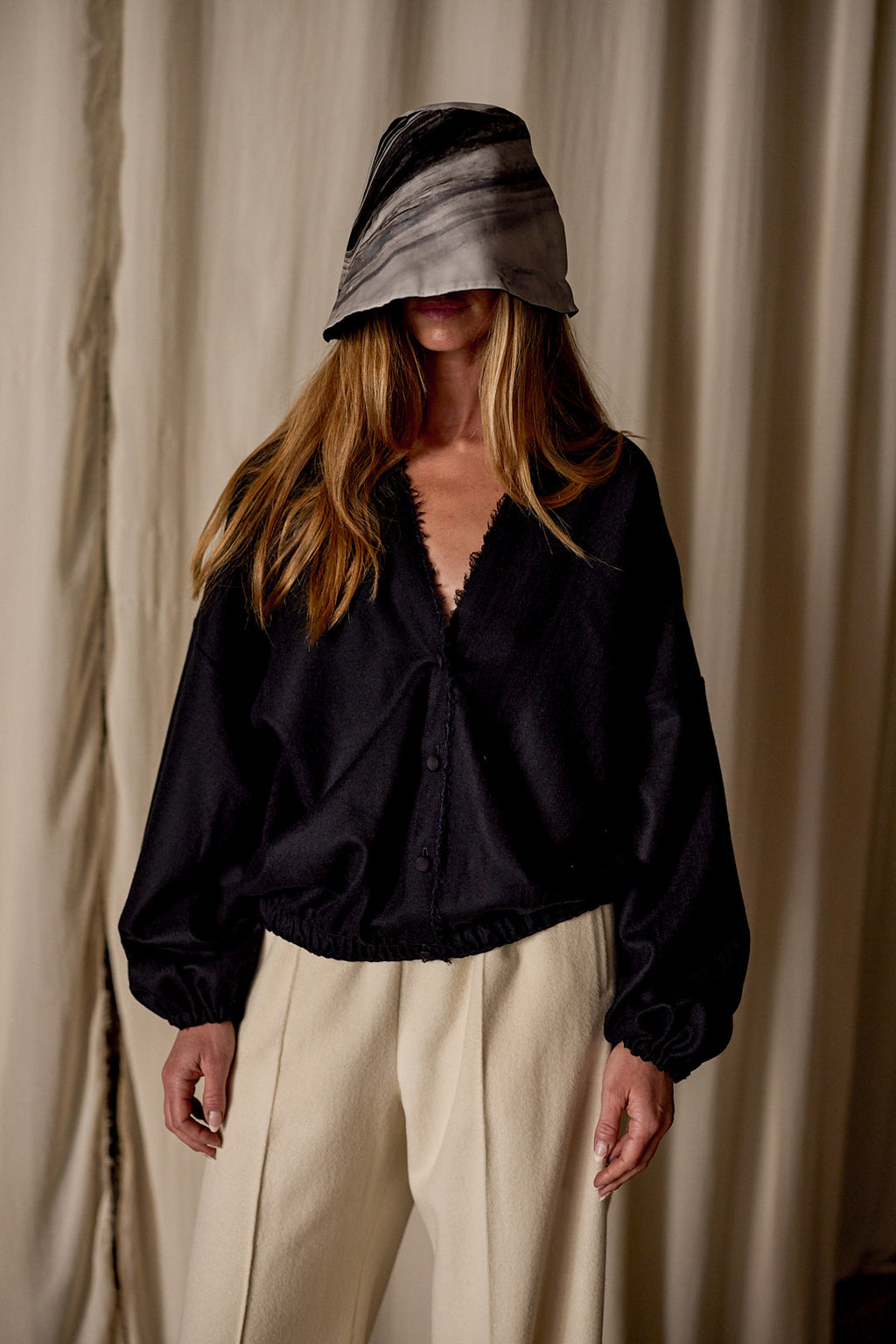 A person with long hair is wearing a wide-brimmed hat that shades their face, a Noir de Chine, and beige pants. The background features light-colored curtains.