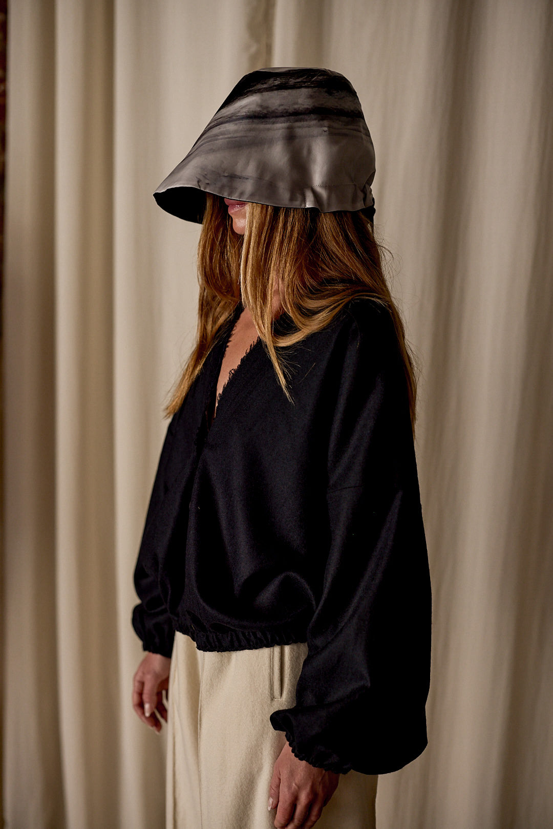 A person with long hair wears a wide-brimmed, tie-dye bucket hat that shadows their face. They are dressed in a loose-fitting black top and light beige pants, complemented by Noir de Chine. The scene is set against a beige curtain backdrop.