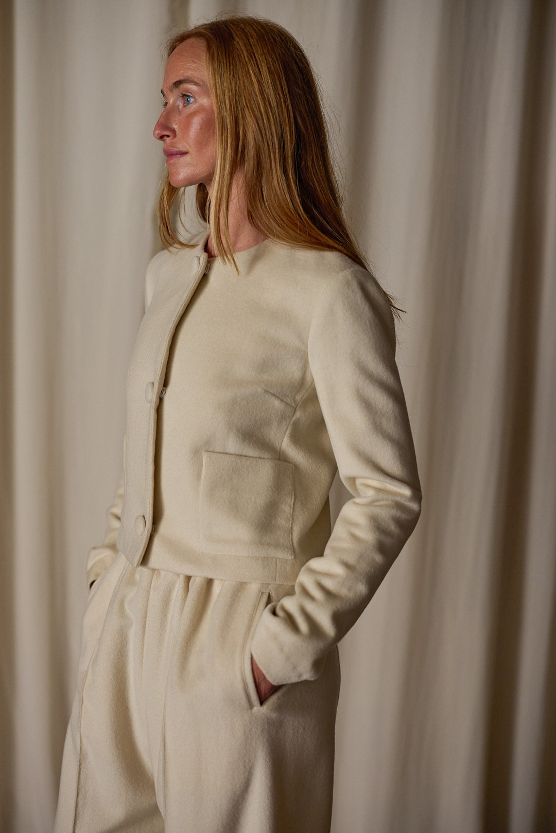 A person with long hair is standing against a plain backdrop, wearing a Crop Cashmere Jacket | Undyed with matching pants. This versatile wardrobe piece, handmade in-house, features light-colored long sleeves and buttoned details. They have their hands in their pockets and are looking to the side.