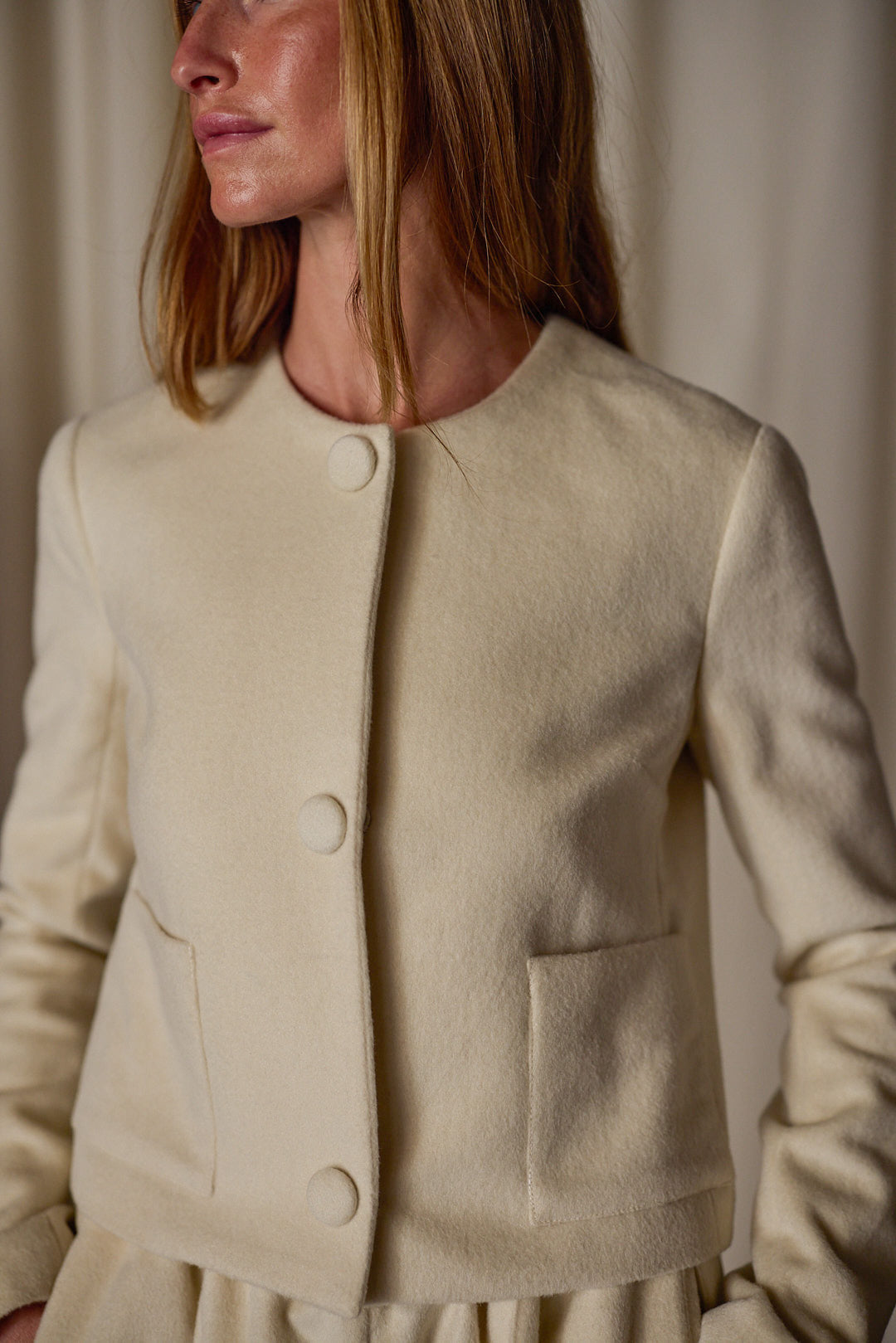 A person with long light brown hair wearing a cream-colored, button-up jacket with two front pockets stands against a neutral background. This versatile wardrobe piece is simple in design with fabric-covered buttons and a round neckline. The Crop Cashmere Jacket | Undyed is handmade in-house.