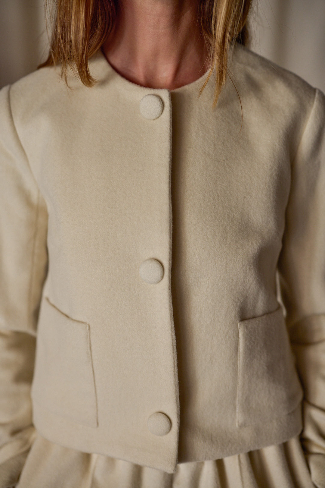 A woman wearing a Crop Cashmere Jacket | Undyed with two front pockets. The jacket has a round neckline and is made of textured fabric. This versatile wardrobe piece focuses on the upper part of her body, from the shoulders to the waist.