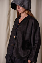 Load image into Gallery viewer, A woman stands wearing a stylish black outfit, featuring a loose-fitting Sumi Top | Black Xiang Yun Silk with gold buttons and large pockets. She also wears a checkered, wide-brimmed hat that shades her face and has her hands in her pockets. The background is a neutral, light-colored fabric highlighting the sustainable design from Xiang Yun Silk.