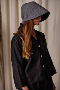 A person with long, light brown hair wears a wide-brimmed gray hat that shades their face. They are dressed in a Sumi Top | Black Xiang Yun Silk with loose sleeves. The background is out of focus, featuring neutral-colored curtains.