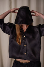 Load image into Gallery viewer, A person with long hair wears a black silk satin Sumi Top | Black Satin Organza, including buttons and a hood that covers most of their face. They have their hands raised, adjusting the hood. The background features a light-colored curtain.