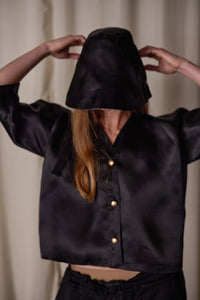 A person with long hair wears a black silk satin Sumi Top | Black Satin Organza, including buttons and a hood that covers most of their face. They have their hands raised, adjusting the hood. The background features a light-colored curtain.