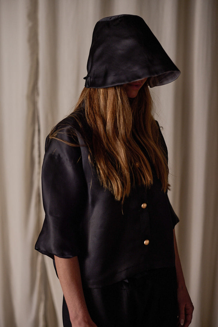 A person with long hair, standing against a draped curtain background, is clad in a black Sumi Top | Black Satin Organza made from sustainable design. The hooded top, featuring two buttons, partially obscures their face.