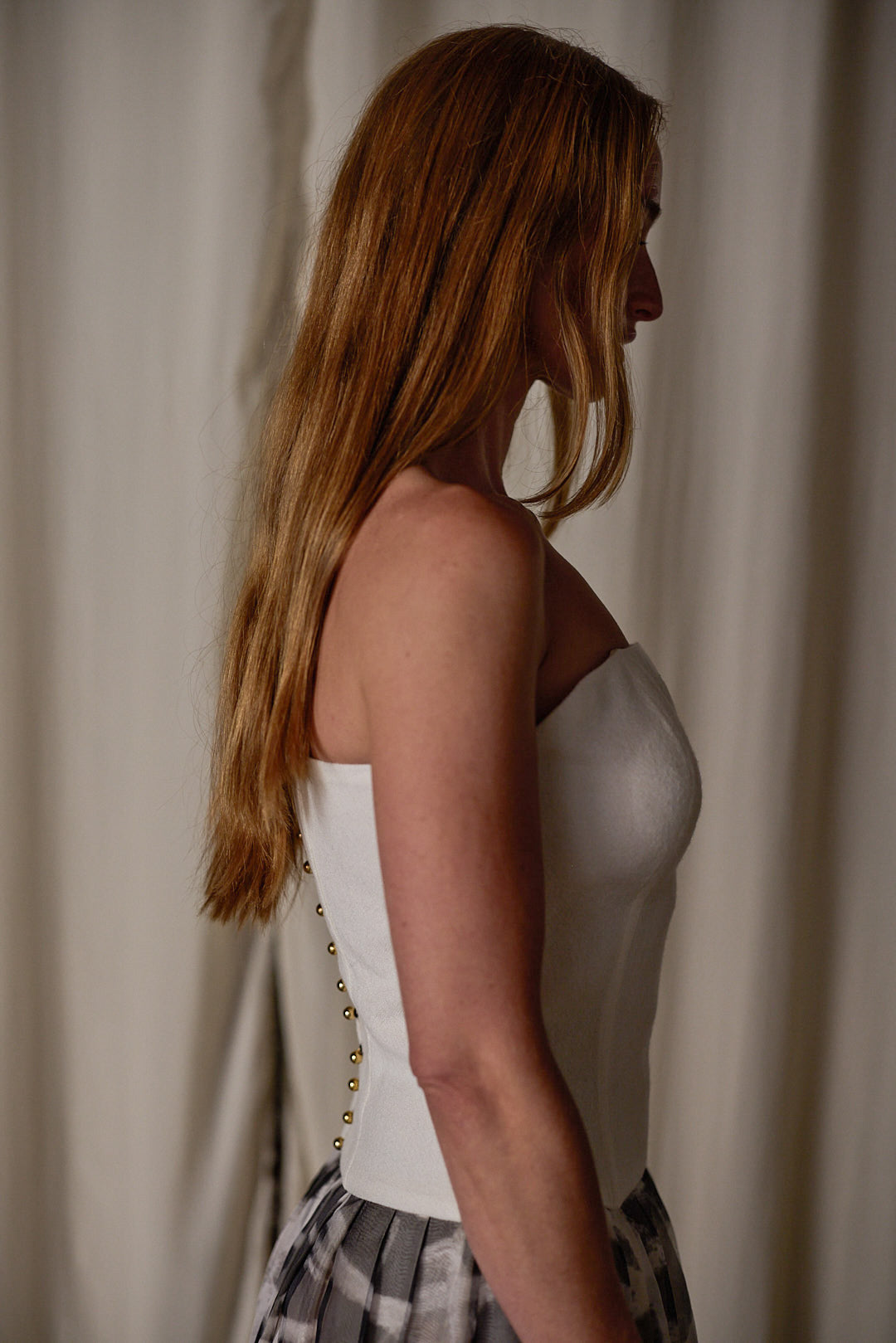 A woman with long blonde hair is shown in profile against a neutral background. She is wearing a Custom Bustier | Japanese Wool Gauze Undyed from Atelier Lan Jaenicke, featuring visible button detailing down the back, and a plaid skirt made of Japanese wool gauze. The setting appears to be indoors with a soft, muted curtain backdrop.