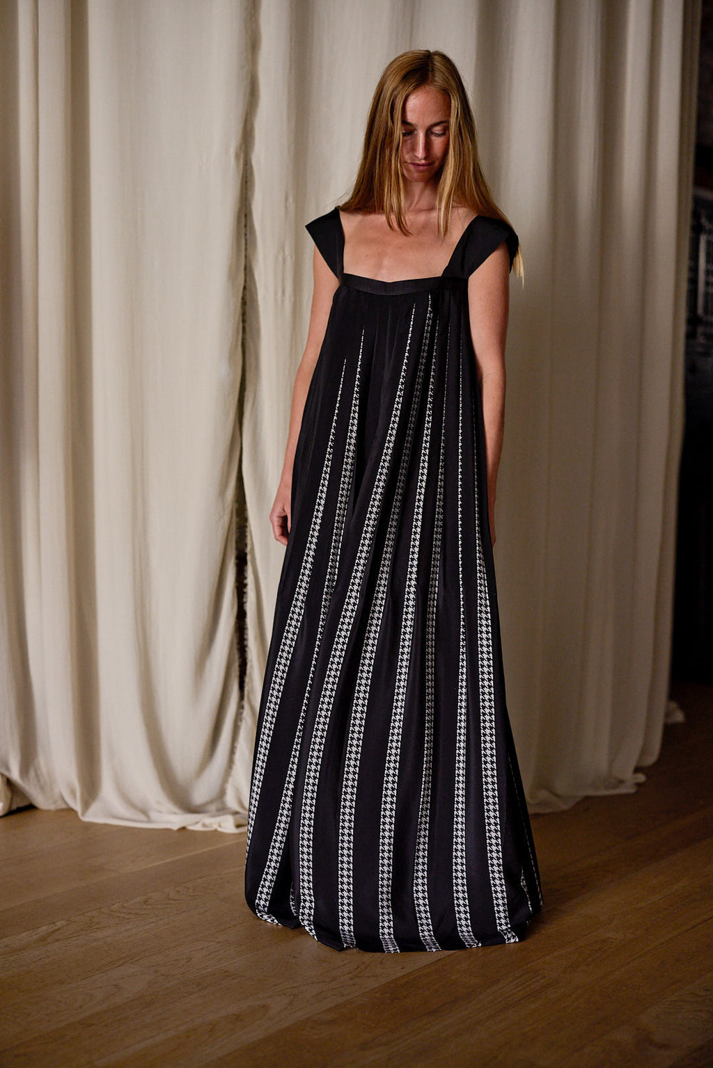 A woman wearing a Hand Pleated Peony Dress Reversible | Custom Houndstooth Stripe featuring thin white vertical stripes stands in front of light-colored, slightly open curtains. She has long blonde hair and is looking down, holding her dress slightly with one hand. The floor is made of light wood.