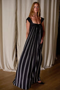 A woman with long hair stands indoors in front of white curtains. She is wearing a flowy, sleeveless black Hand Pleated Peony Dress Reversible | Custom Houndstooth Stripe adorned with white vertical stripes and a wide square neckline. Her hands rest in the dress's pockets, and she is looking confidently at the camera.