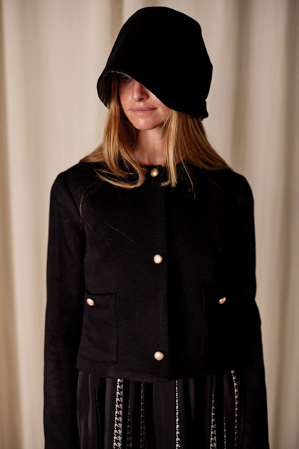 A person with long, blonde hair wears a black hooded hat, obscuring part of their face. They are dressed in a Crop Cashmere Jacket | Black with gold buttons and a black skirt with white stitching details. The background is a simple, light-colored curtain.