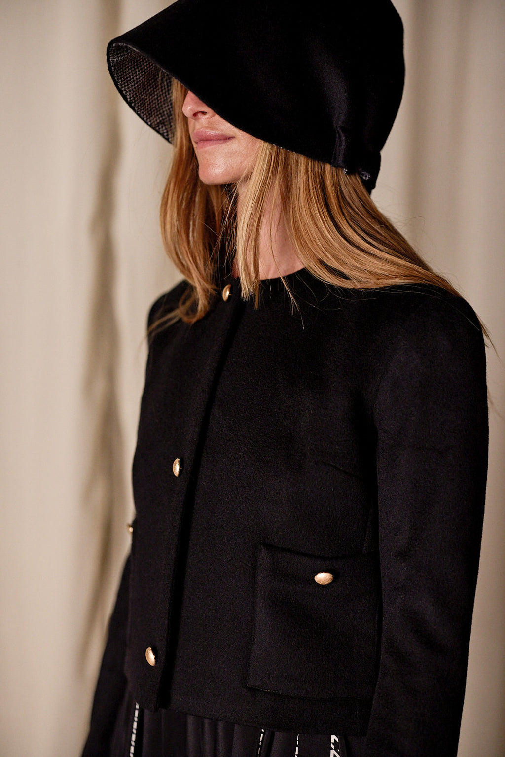 A woman with long hair stands against a neutral background, wearing the Crop Cashmere Jacket | Black with gold buttons and a large black hat that shades her eyes. Handmade in San Francisco, the jacket features two front pockets, and the overall look is minimalist and stylish.