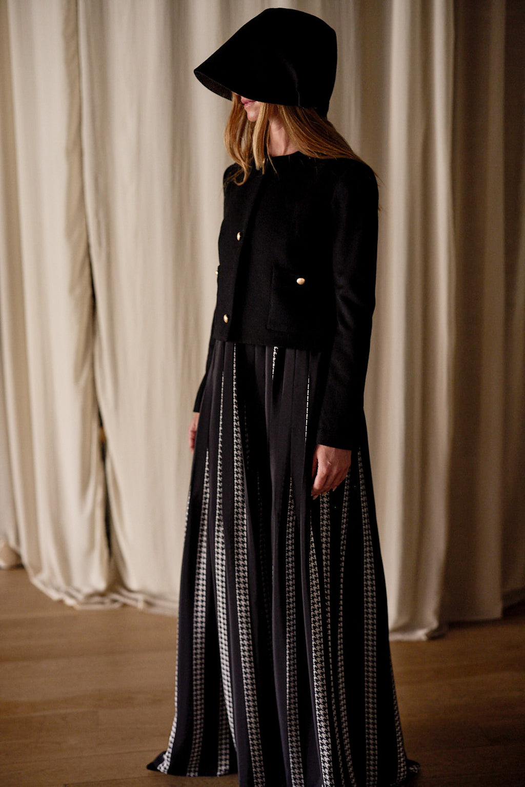 A person is standing indoors, dressed in a long, black and white vertical striped skirt, paired with a handmade Crop Cashmere Jacket | Black adorned with gold buttons. Their head is covered with a black hat casting a shadow over their face. The background features light, pleated curtains.