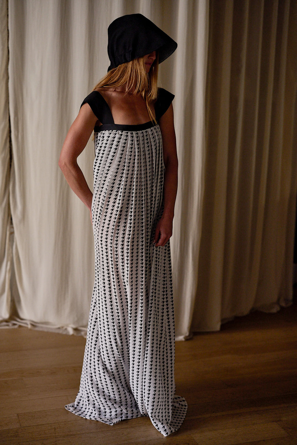 A person stands indoors against floor-length white curtains, elegantly clad in a loose, floor-length Hand Pleated Peony Dress | Crepe de Chine Custom Houndstooth made of silk crepe de chine with a black and white dotted pattern. Their face is partially obscured by a dark hood attached to the dress. The setting has wooden flooring.