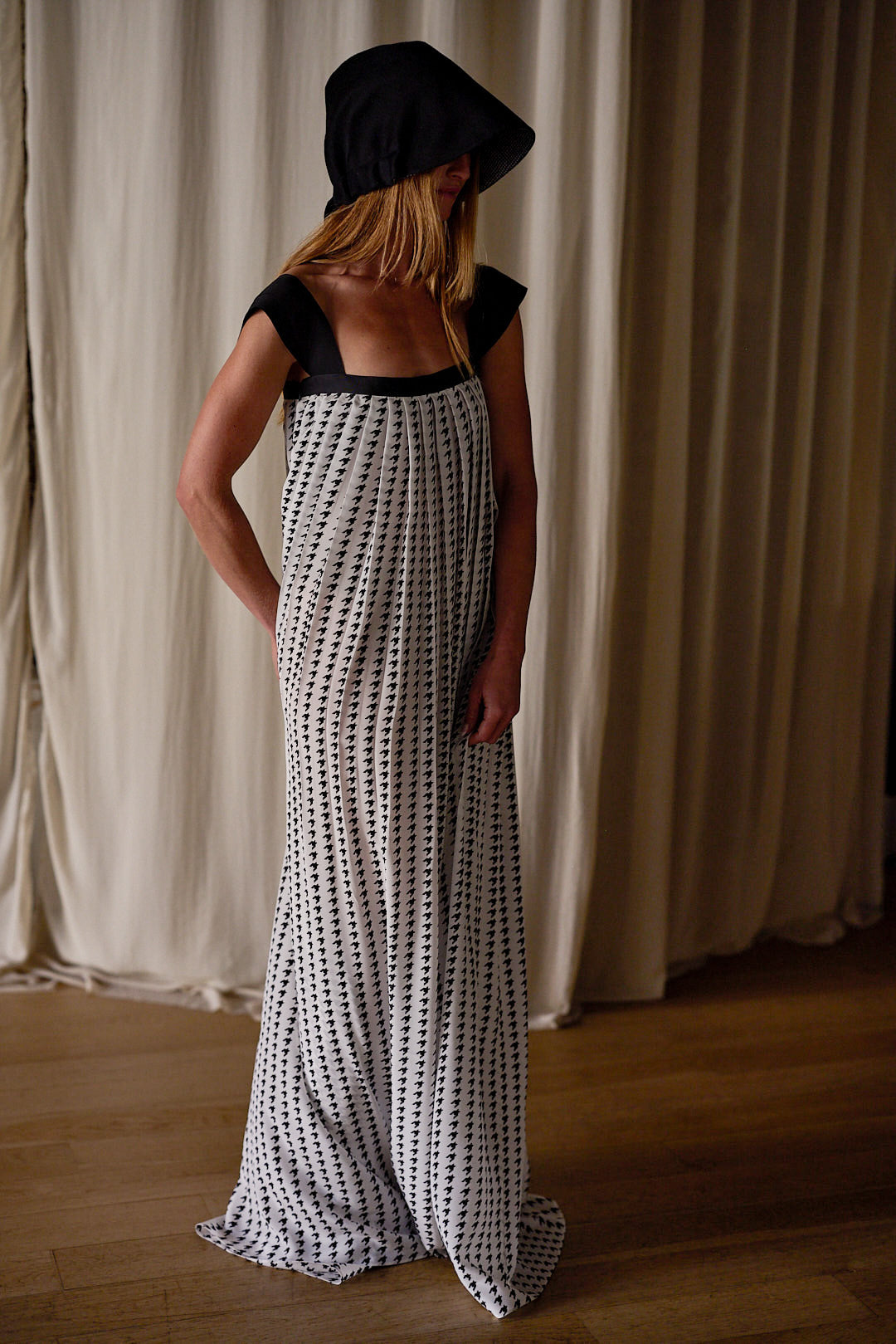 A person stands indoors against floor-length white curtains, elegantly clad in a loose, floor-length Hand Pleated Peony Dress | Crepe de Chine Custom Houndstooth made of silk crepe de chine with a black and white dotted pattern. Their face is partially obscured by a dark hood attached to the dress. The setting has wooden flooring.