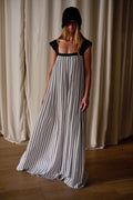 A person stands indoors wearing a long, flowy Hand Pleated Peony Dress | Crepe de Chine Custom Houndstooth with black patterns. The dress has black shoulder straps and a square neckline. They also have on a black hat that partially obscures their face. The background features white curtains and a wooden floor, with an air of zero waste design elegance.