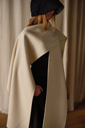 Person wearing a stylish, cream-colored Doubleface Cashmere X-Cape over a black outfit with a black wide-brimmed hat covering their face. The background features neutral, draped curtains.