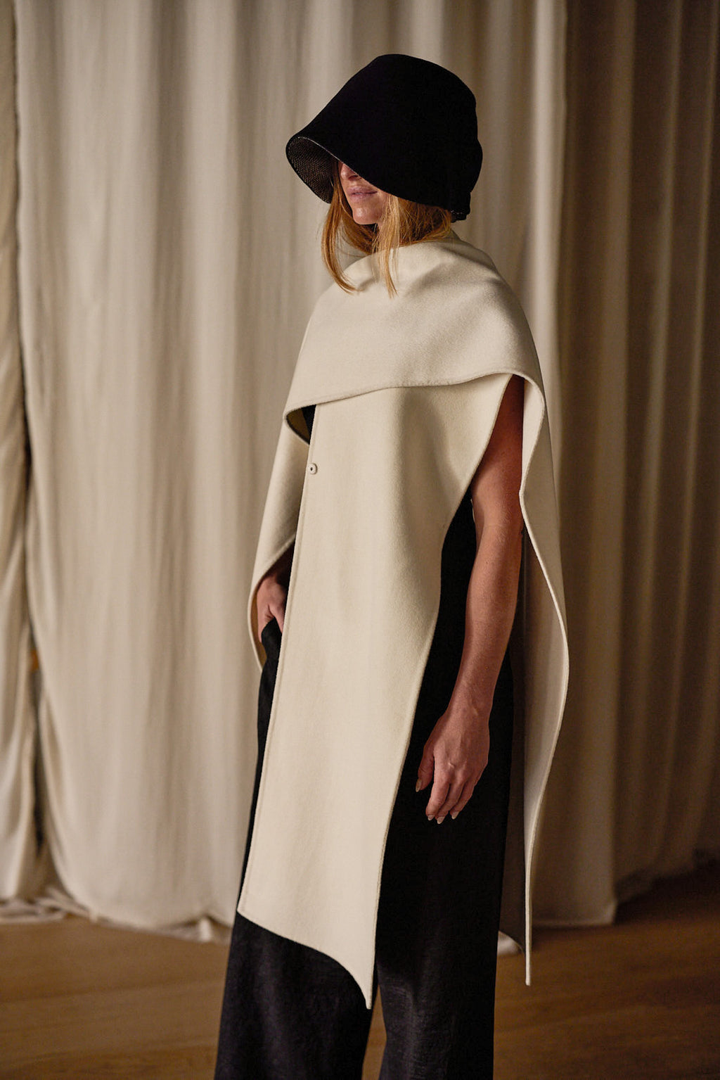 A person wearing a black wide-brimmed hat and a stylish off-white cape stands against a backdrop of beige curtains. The Doubleface Cashmere X-Cape drapes elegantly over their shoulders, handmade in-house for quality, offering multiple ways of styling as they gaze downward.