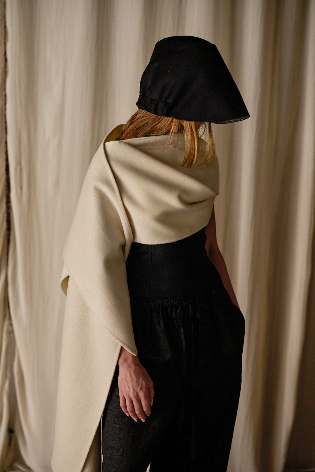 A person stands with their back to the camera, wearing a black hat and an off-white Doubleface Cashmere X-Cape crafted from 100% double-faced woven cashmere, paired with black pants. The beige curtain backdrop highlights the fashion-forward and minimalist design, showcasing multiple ways of styling the luxurious piece.