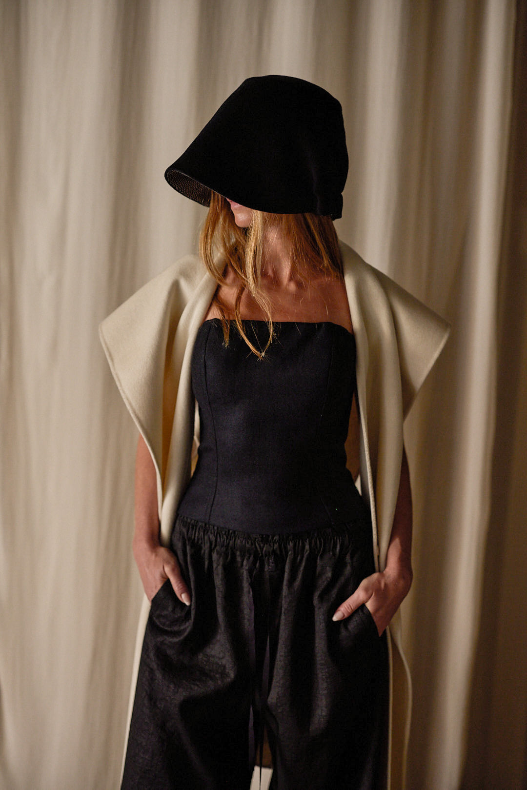 A woman stands against a neutral-colored backdrop. She is wearing a black, wide-brimmed hat that partially obscures her face, a strapless black top, loose black pants, and a Doubleface Cashmere X-Cape draped over her shoulders. Her hands are in her pockets.