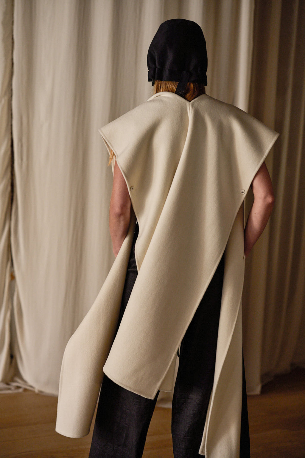 A person stands facing away, wearing a Doubleface Cashmere X-Cape made from 100% double-faced woven cashmere that drapes down their sides, over a black outfit. They are also wearing a black head covering. The background consists of beige curtains. The coat offers multiple ways of styling.