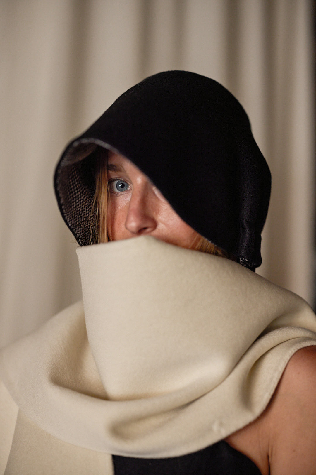 A person with blue eyes is partially hidden under a large black hood and a Doubleface Cashmere X-Cape, that covers their neck and lower face. The handmade in-house accessory offers multiple ways of styling. The background is out of focus.