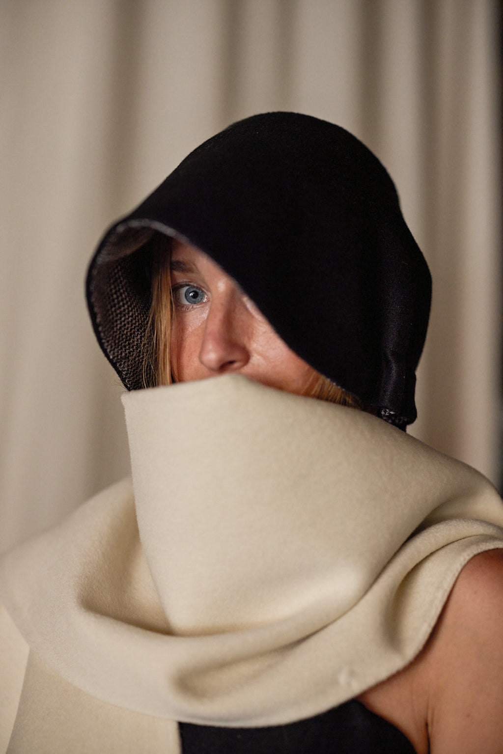 A person with light-colored hair partially hidden wears a black hood that shadows their face and a large, cream-colored Cashmere Ribbon Cloche | Black, Reversible covering their mouth. The background is blurred and neutral-toned.