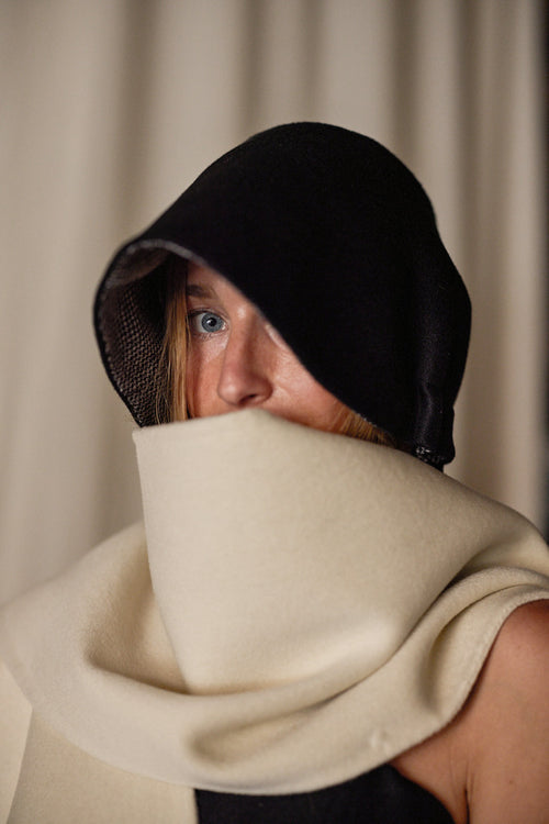A person with light-colored hair partially hidden wears a black hood that shadows their face and a large, cream-colored Cashmere Ribbon Cloche | Black, Reversible covering their mouth. The background is blurred and neutral-toned.
