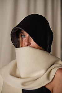 A person with light-colored hair partially hidden wears a black hood that shadows their face and a large, cream-colored Cashmere Ribbon Cloche | Black, Reversible covering their mouth. The background is blurred and neutral-toned.