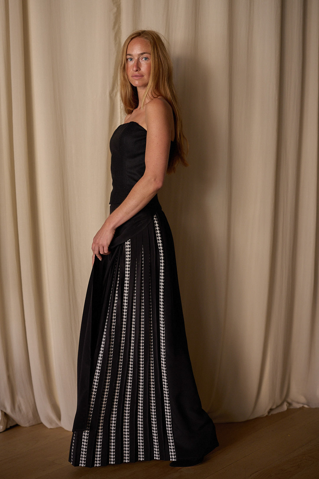 A woman with long hair is wearing an elegant, strapless black Layered Skirt w/ Hand Pleats | Crepe de Chine adorned with vertical white detailing on the skirt. The custom houndstooth stripe adds a unique touch. She is standing sideways, facing slightly towards the camera, with a neutral expression. The background features soft, cream-colored drapes.
