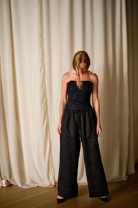 A woman with long brown hair stands indoors against a backdrop of cream-colored curtains. She is wearing a black strapless top and high-waisted, wide-leg Xiang Yun Silk Trousers. She looks downwards with her hair slightly covering her face.