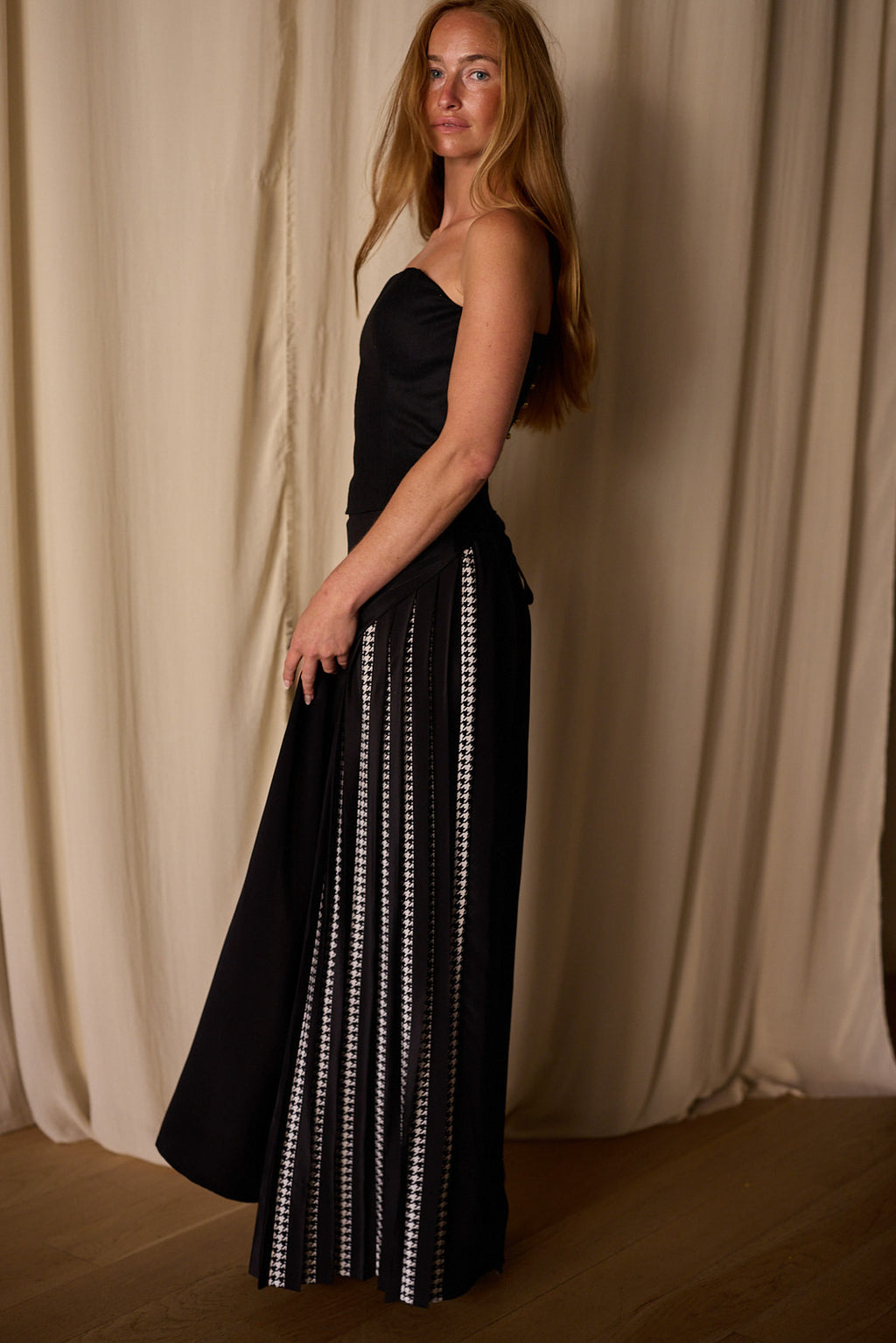 A woman with long, straight hair stands in front of beige curtains. She is wearing a strapless black dress featuring custom houndstooth stripes on the lower half. The Layered Skirt w/ Hand Pleats | Crepe de Chine is fitted at the top and flows loosely from the waist down. She gazes into the camera with an air of confidence.
