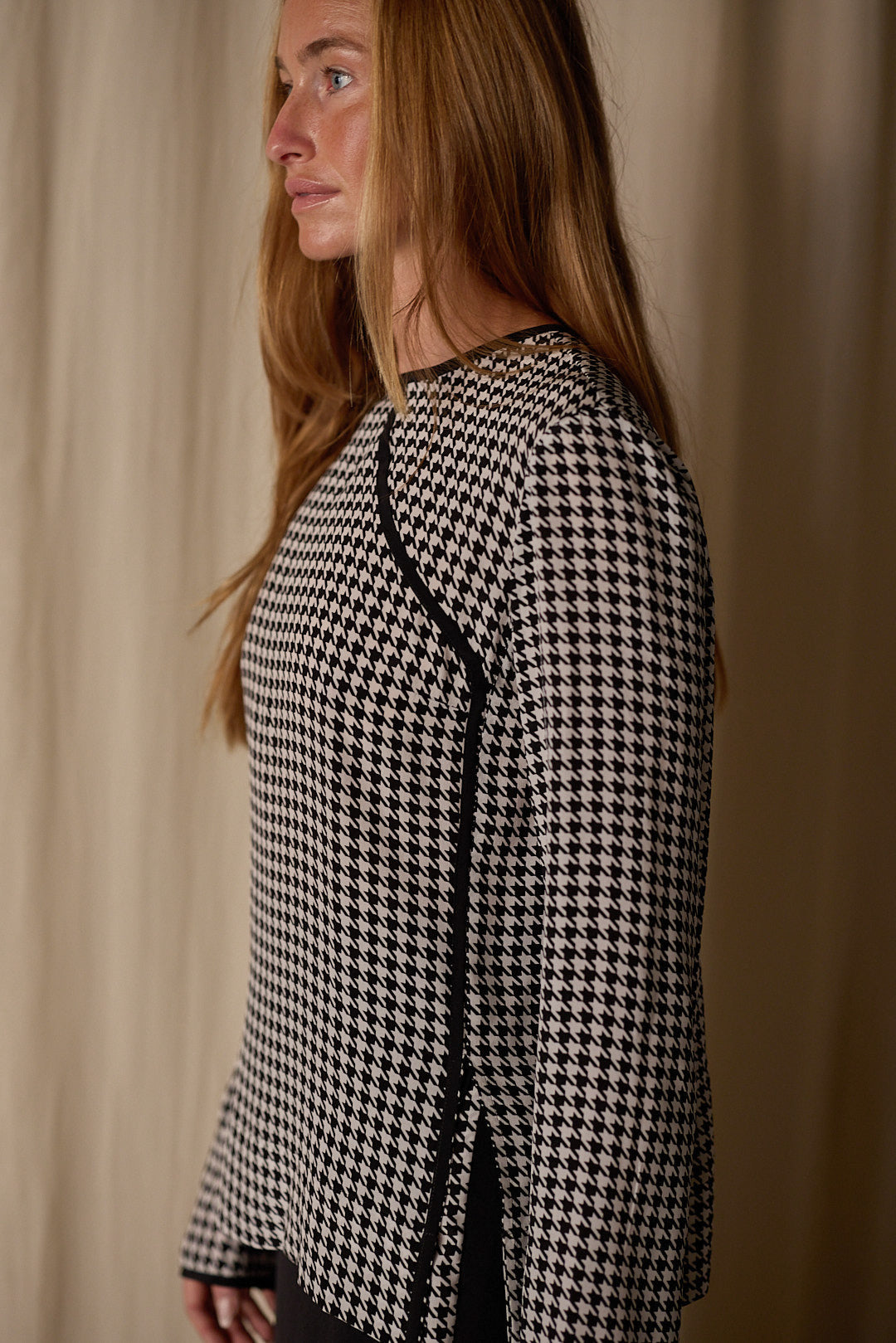 A person with long, straight hair is shown in profile, wearing a black and white custom Qipao Blouse | Crepe de Chine Houndstooth with black trim. They are standing against a beige, textured background.